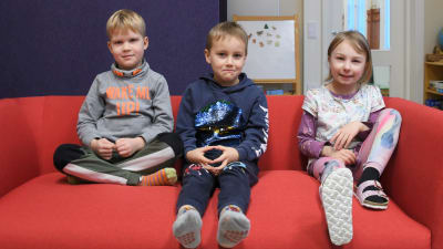 Bilden föreställer tre glada förskolebarn som sitter i en röd soffa.