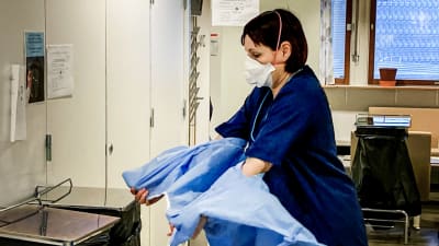 Sjukskötare Sanna Nerjanto tar av sig en ljusblå skyddsjacka, ovanpå munnen och näsan har hon ett munskydd.