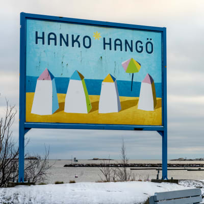 Hangö-skylt vid en strand. På skylten finns fyra ritade badhytter, sol, strand, blå himmel och texten Hanko-Hangö.