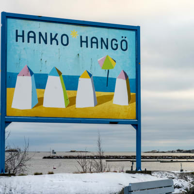 Hangö-skylt vid en strand. På skylten finns fyra ritade badhytter, sol, strand, blå himmel och texten Hanko-Hangö.