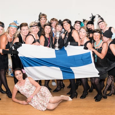 En grupp kvinnor i svarta klänningar står och håller i en pokal och en finsk flagga.