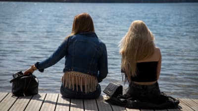 Två tonåriga flickor sitter med ryggen mot kameran på en brygga i sommartid.