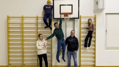 Fyra barn och en vuxen står i en gymnastisal. Tre av barnen har klättrat uppför ribbor som finns vid väggen, en av dem hänger till och med i en basketkorg.