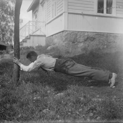 En man lutar mot en trädstam och sträcker ut sig vilande på sina tår. Bilden är tagen 1930 i Bromarf.