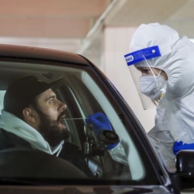 En person i skyddsutrustning tar ett coronatest på en person som sitter i en bil.