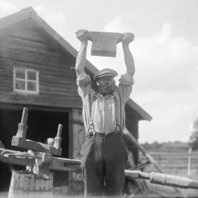En man håller ett smidesstäd ovanför sitt huvud. Bilden är tagen 1930 i Finström, Åland.