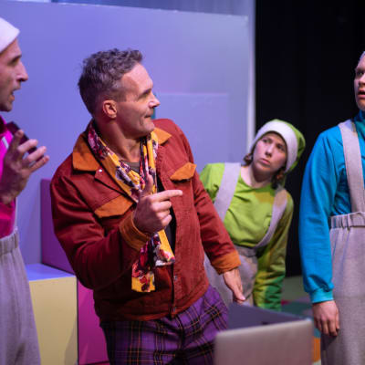Tre skådespelare i pastellfärgade tomtekläder tittar förvånat på en fjärde man i ljusbrun skinnrock mellan dem. 