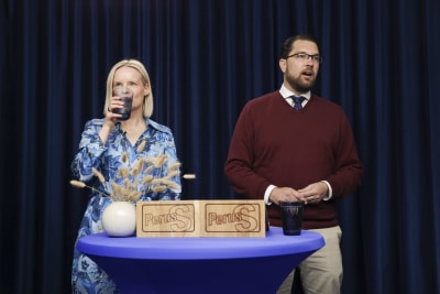 Riikka Purra dricker ett glas vatten medan Jimmie Åkesson talar. De står bakom ett blått bord.