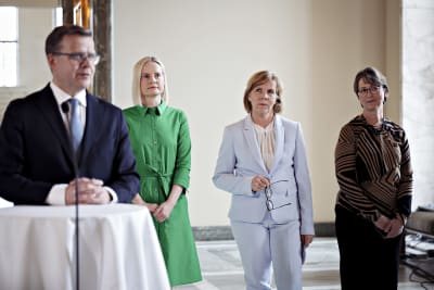 Samlingspartiets ordförande Petteri Orpo står vid en mikrofon på en presskonferens i riksdagen, Sannfinländarnas partiledare Riikka Purra, SFP:s Anna-Maja Henriksson och KD:s Sari Essayah står bakom. 