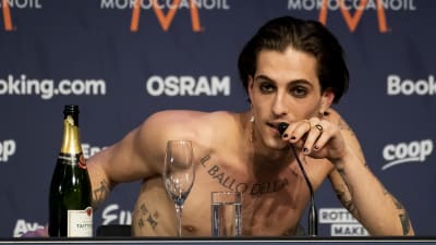 Damiano David, utan skjorta, vid en presskonferens. Han håller i en mikrofon, och har en champagneflaska framför sig.