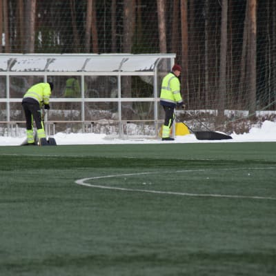 Två män skottar bort litet snö från den annars gröna konstgräsplanen i Karis den 6 mars 2018.