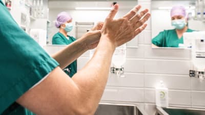 En sjukskötare tvättar händerna i ett badrum. I spegeln syns två sjukskötare som bär ansiktsskydd.