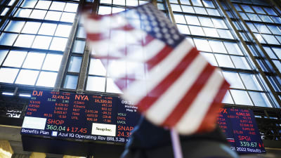 USA:s flagga vajar i förgrunden, i bakgrunden aktiebörser på en elektronisk tavla. Börserna går neråt.