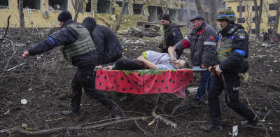 Flera män bär en gravid kvinna på en bår. I bakgrunden ett sönderbombat hus.