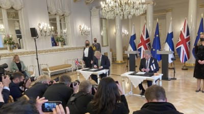 Premiärminister Boris Johnson och president Sauli Niinistö undertecknar papper på Presidentens slott. I förgrunden syns journalister. I bakgrunden syns Finlands, Storbritanniens och EU:s flaggor.