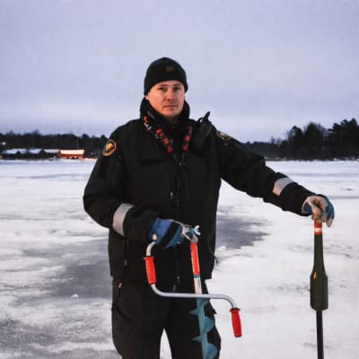 Man i sjöbevakardräkt står på is med en isborr och en mätstick i händerna.