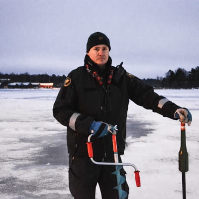 Man i sjöbevakardräkt står på is med en isborr och en mätstick i händerna.