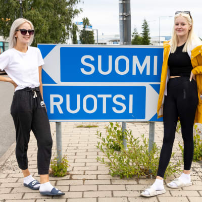 Kaksi nuorta naista poseeraa Suomi-Ruotsi kyltin edessä Torniossa