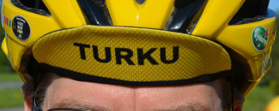 En gul skärmmössa med texten Turku sticker fram under en gul cykelhjälm.