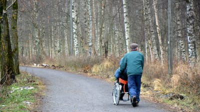 En äldre man kör en person i rullstol längs promenadstråket runt Metviken i Vasa