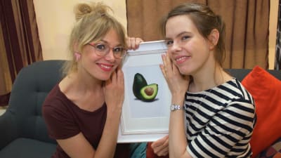 Två leende kvinnor sitter i en soffa med en bild på en avocado mellan sig.