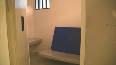 Bild in mot cell i häkte. Bord, brits med blå madrass, litet fönster med galler.