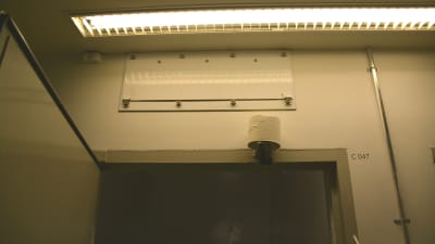 Bild av övre delen av dörröppning till en cell. högst uppe synns ett lysrör. Ovanför dörröppningen hänger en toapappersrulle.
