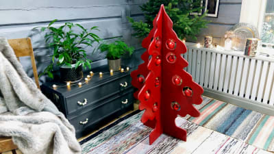 En röd julgran av trä som är monterad av två skivor utformade som en gransiluett. I bakgrunden en liten julgran. Julgranarna är fotograferade i ett vardagsrum med en svart byrå i bakgrunden och trasmattor på golvet. den röda julgranen är dekorerad med röd