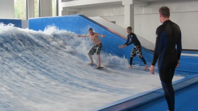 Två personer surfar i en inomhus surfbassäng medan en tittar på.