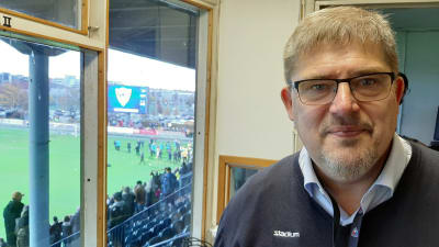 Rainer Svens, en man grånande hår, grånande skägg och glasögon. I bakgrunden firar fotbollsspelare nere på planen att de vunnit.
