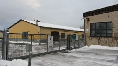 Två fabriksbyggnader och ett staket av metallnät. Stängd port till området. Snö och vinter.