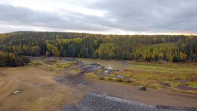 En våtmark anläggs i Huskvarn, Snappertuna, Raseborg. Drönarbild visar grävmaskiner och dumpers i höstligt landskap, i förgrunden ett vattenområde och åkrar, i bakgrunden skog. 