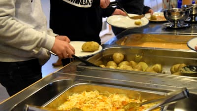 Några personer tar åt sig mat från en buffé där det finns råkostsallad, potatis och kycklingsås.