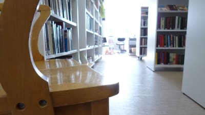 En gammal trästol i en ny ljus biblioteksmiljö. Vita hyllor fyllda med böcker. Ljus strömmar in genom ett fönster.