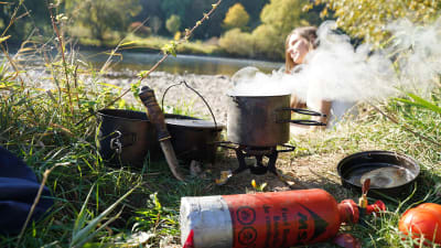 Matlagning ute i naturen. I förgrunden syns en liten bensinflaska som är kopplad till ett sorts spritkök. Ovanpå ligger en kastrull som det ryker ur. I bakgrunden syns en kvinna.