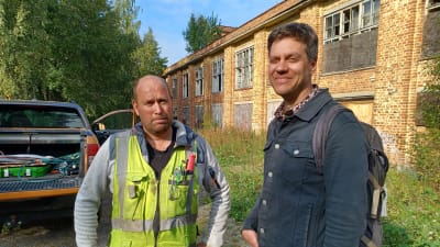 Två män, en i gul arbetsväst och en tjänsteman med ryggsäck på står framför en gul tegelbyggnad som liknar en ruin.