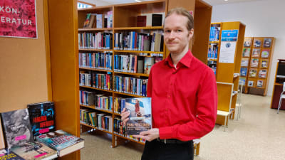 En man står och håller fram en bok i ett bibliotek.