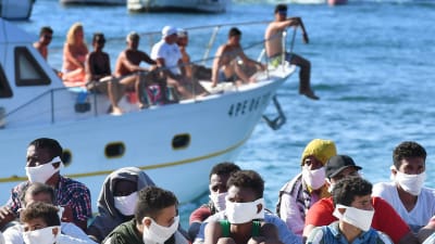 Nyligen anlända migranter satt uppradade på kajen i Lampedusa på onsdagen, medan en turisbåt med italienska turister passerade.
