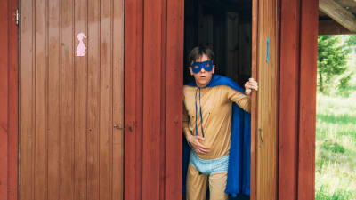 Supersankariasuun pukeutunut mies tulee ulos ulkokäymälästä vatsaansa pidellen.