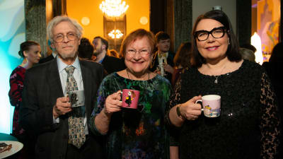 Presidentti Tarja Halonen, Pentti Arajärvi ja Ylen toimitusjohtaja Merja Ylä-Anttila hymeilevät kameralle muumimukit käsissään.