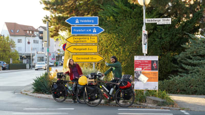 Två cyklister står framför några gula vägskyltar i Tyskland. Deras cyklar är fullastade med väskor. De ser glada ut. Den ena pekar på skyltarna.