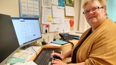 En kvinna sitter vid sitt skrivbord och tittar in i kameran. 