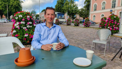 En man i blå skjorta sitter och dricker kaffe vid ett bord på ett torg.