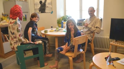 En familj sitter runt ett matbord. Vid första anblicken ser det ut att vara bara en man, en ung flicka och en pojke, men när man tittar närmare ser man att stolen pojken sitter på egentligen är en människa utklädd till stol.
