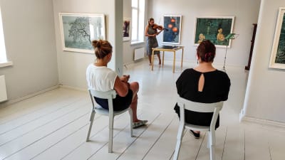 Två kvinnor sitter på varsin stol inne i ett vitmålat rum och ser på en kvinna som spelar violin.