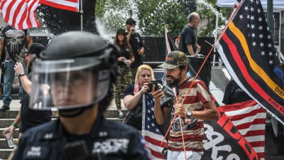 En polis i skyddsutrustning står i förgrunden, i bakgrunden demonstranter som håller i amerikanska flaggan.
