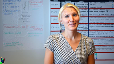 Marina Kinnunen står i sitt kontor. Hon bär en rutig klänning och i bakgrunden syns en whiteboardtavla.