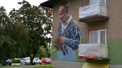 En äldre man i blå skjorta har målats på väggen. Han håller en bok med pip i sin hand.