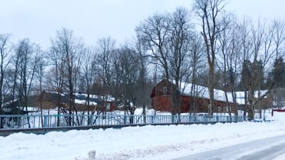 En vintrig vy över ett område med gamla byggnader, mycket snö, bilden tagen från en bro, vägen och broräcket syns i förgrunden. Bara träd.