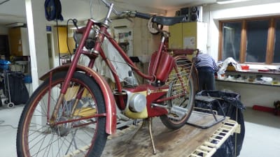 En gammal röd moped på ett bord i en verkstad. Så gammal moped att den mer liknar en cykel